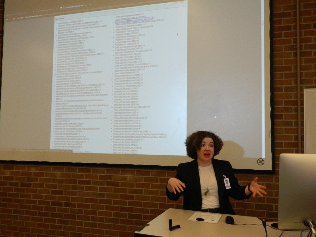 A person with curly hair midsentence with a database projected on to the screen behind them.